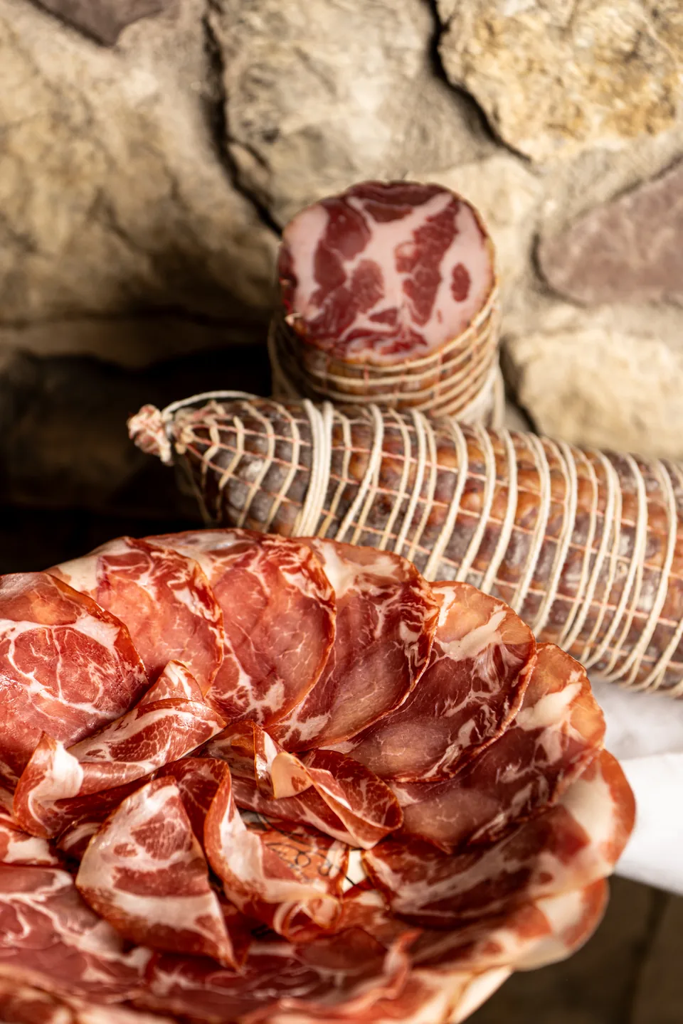 Capocollo intero e affettato su un piatto, davanti a una parete in pietra.